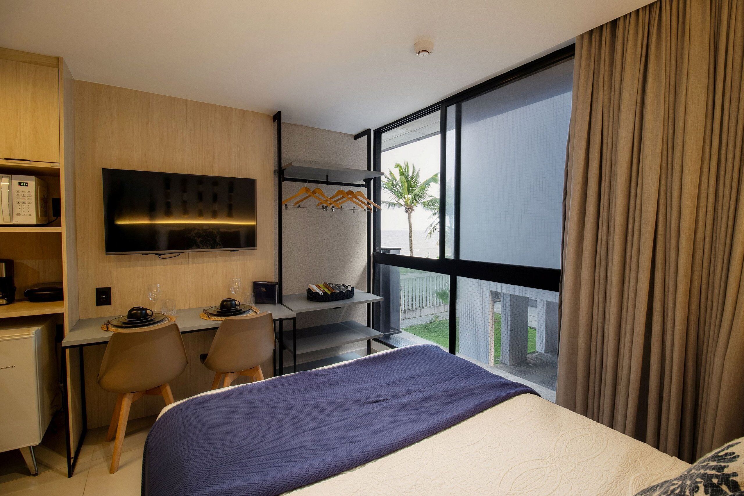 Una camera d'hotel con un letto, una scrivania, sedie, una TV e un'ampia finestra con tende.