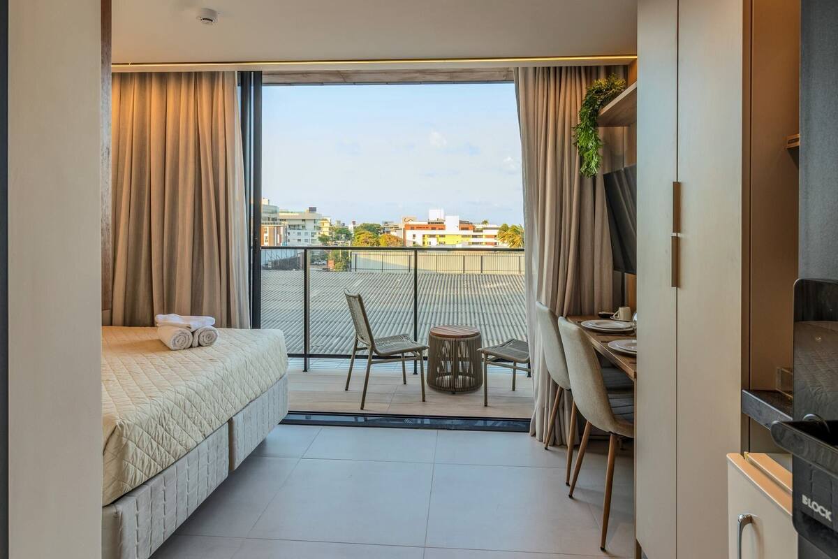 A small, modern bedroom with a bed, a desk, and a chair, a balcony with a view of the city, and a TV.