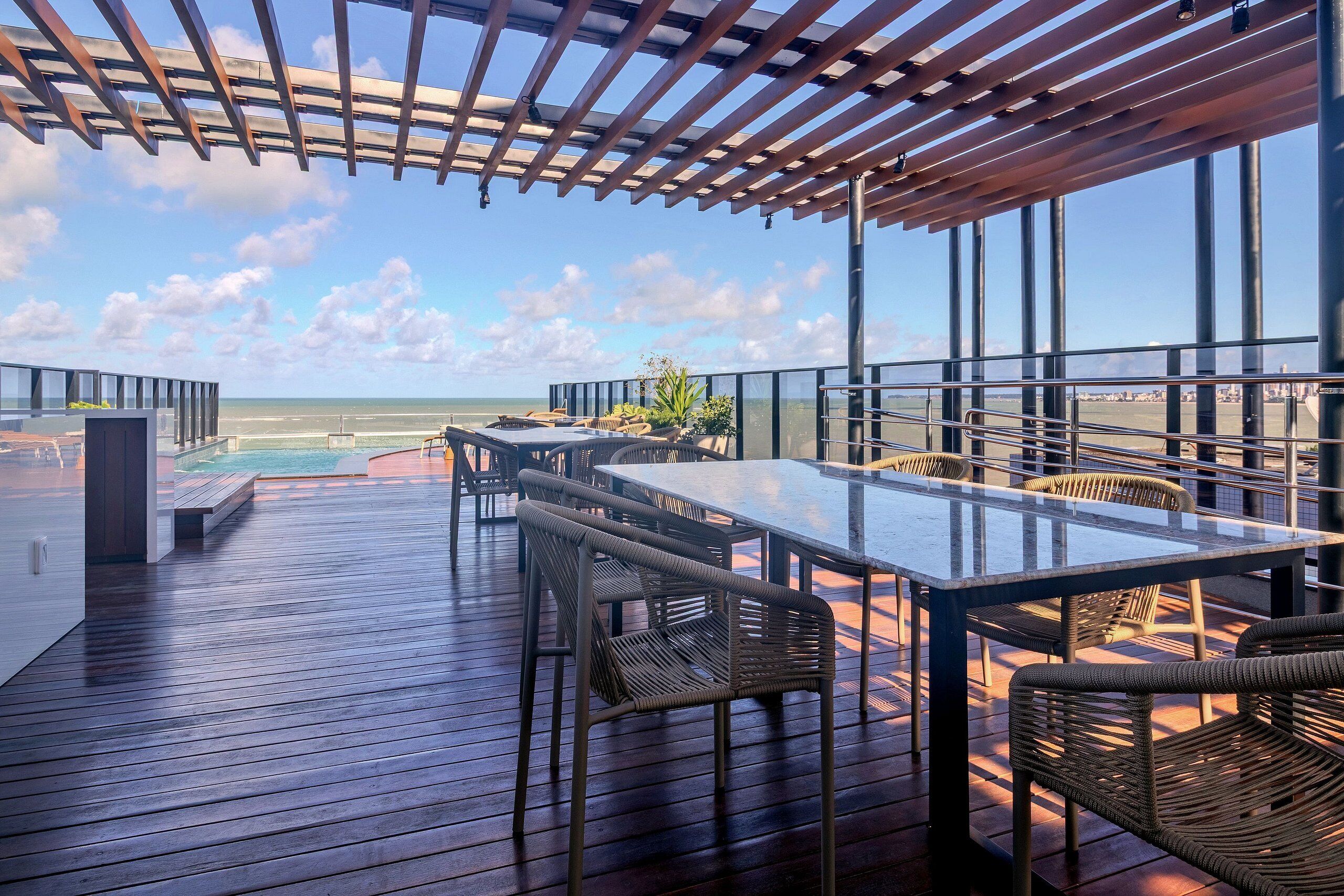 A wooden deck with a glass table and wicker chairs, overlooking a pool and beach.