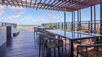 A wooden deck with a glass table and wicker chairs, overlooking a pool and beach.