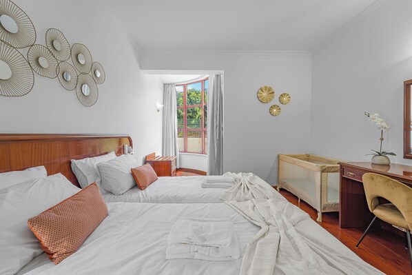 A hotel room with two beds, a desk, a chair, and a window with a view of greenery.