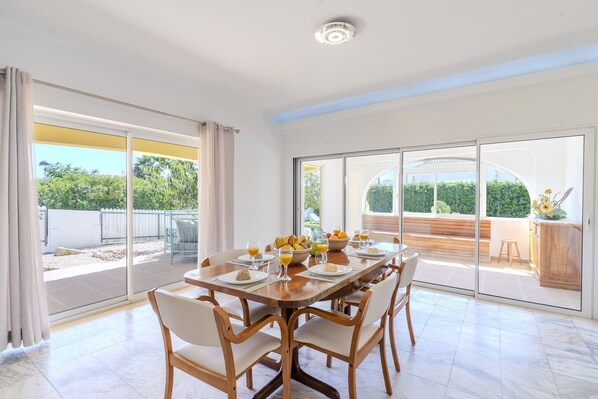 A dining area with a large table set for six, a glass door leading to a balcony, and a view of greenery outside.