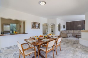A dining area with a wooden table set for four, surrounded by chairs. A kitchen with a refrigerator and a fruit bowl is visible in the background.
