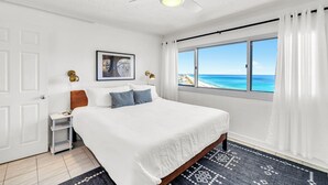 A bedroom with a bed, a nightstand, a ceiling fan, a window with a view of the ocean, and a framed picture on the wall.
