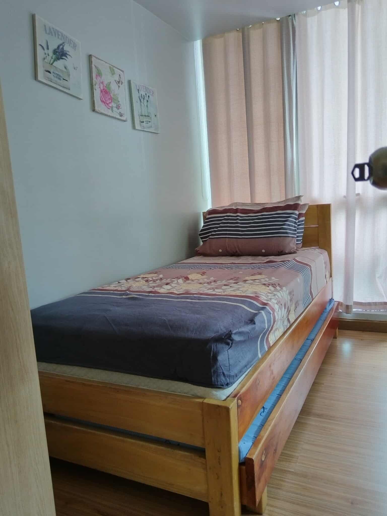 A single bed with a wooden frame, a floral bedspread, and a window with sheer curtains.