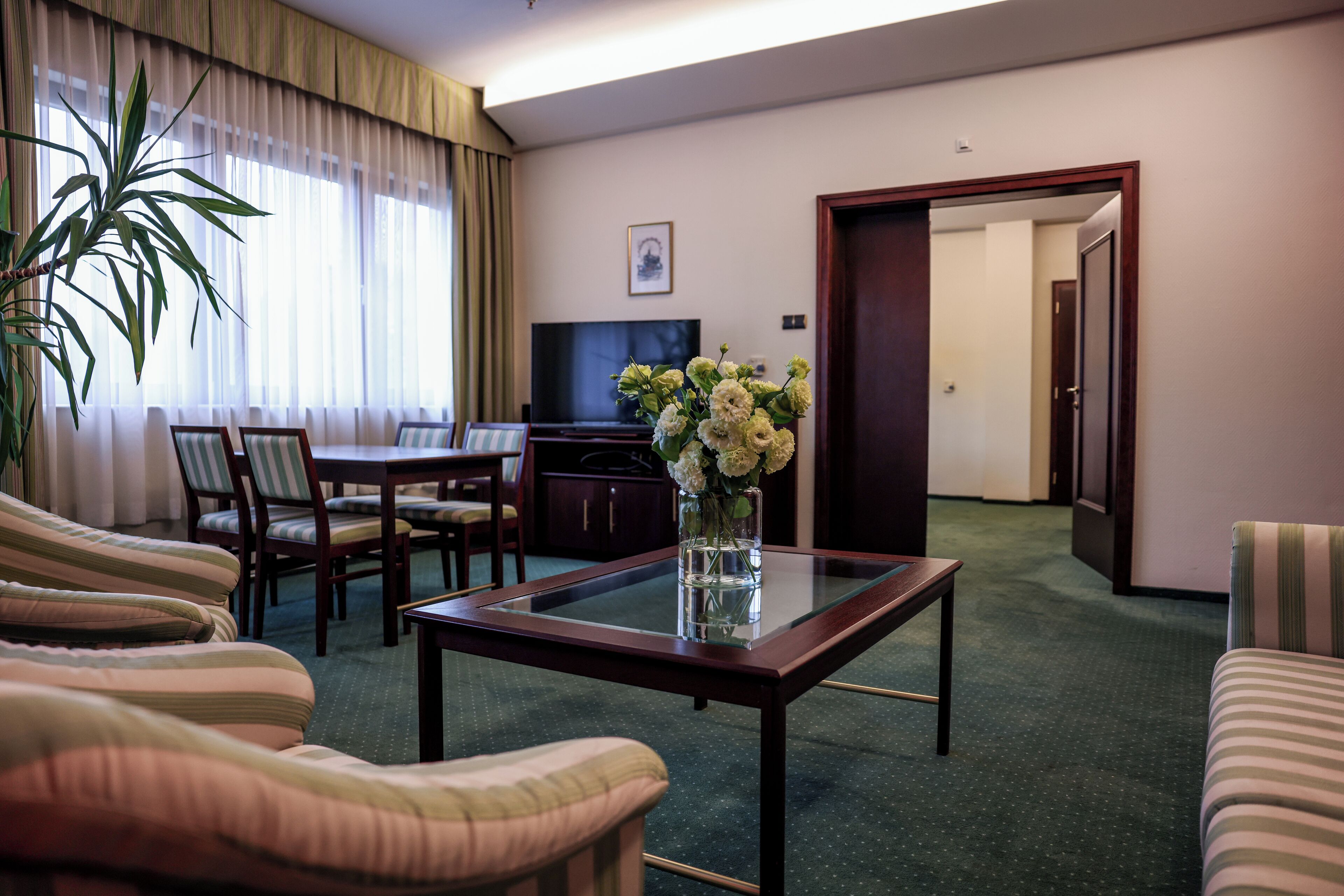 A hotel room with a dining area, a TV, a vase with flowers, and a large window with curtains.