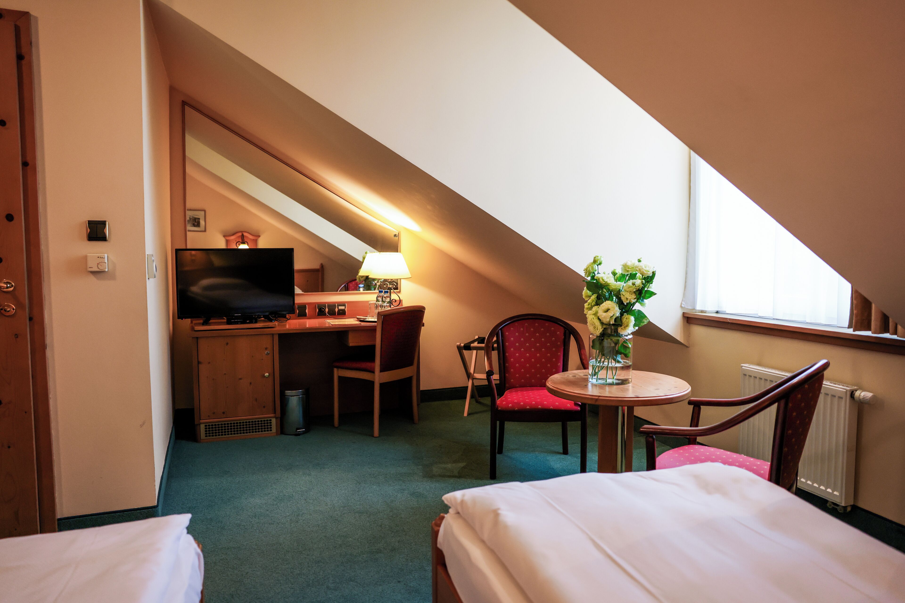 Une chambre d’hôtel avec un lit, un bureau avec une télévision, une chaise, une petite table ornée d’un vase de fleurs et une fenêtre donnant sur un beau paysage.