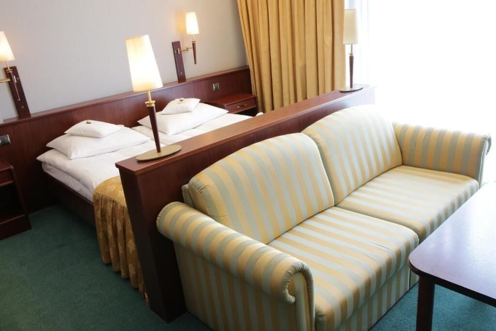 Una habitación de hotel con una cama, un sofá a rayas, una mesita de noche de madera y dos lámparas de noche.