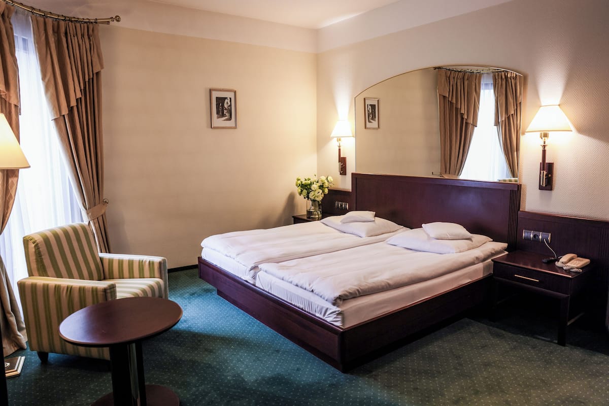 A hotel room with a double bed, a chair, a small table, a nightstand with a lamp, and a mirror.