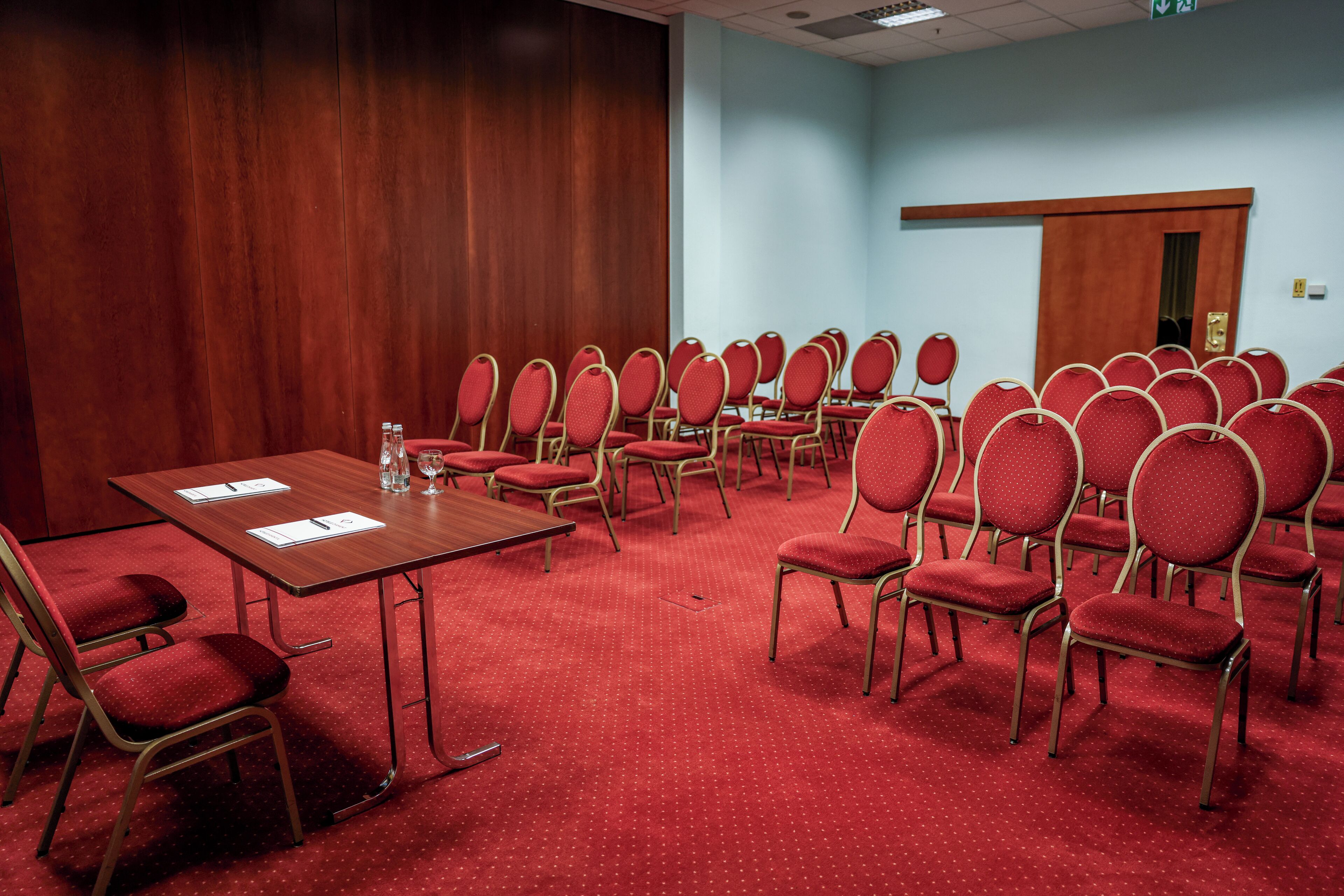 Un salón de conferencias con una larga mesa y filas de sillas rojas, un panel de pared de madera y una pared azul claro con una puerta.