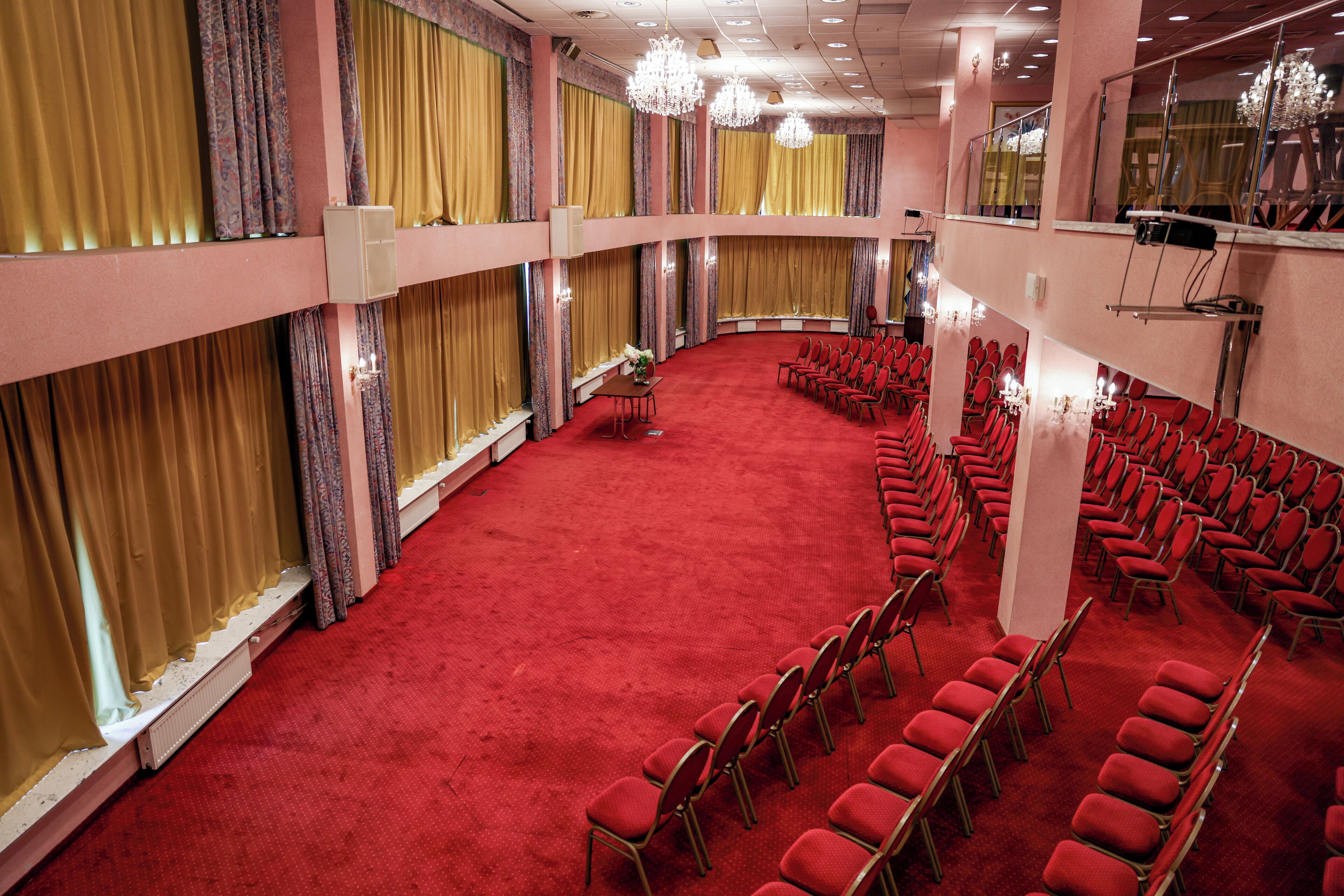 A large hall with rows of red chairs, a red carpet, and golden curtains.