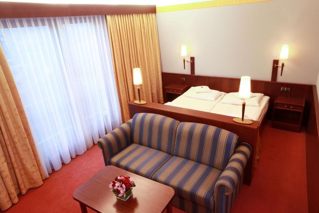 Et hotelværelse med en seng, to sengeborde med lamper, en stribet sofa, et lille bord med en vase blomster og et vindue med gardiner.