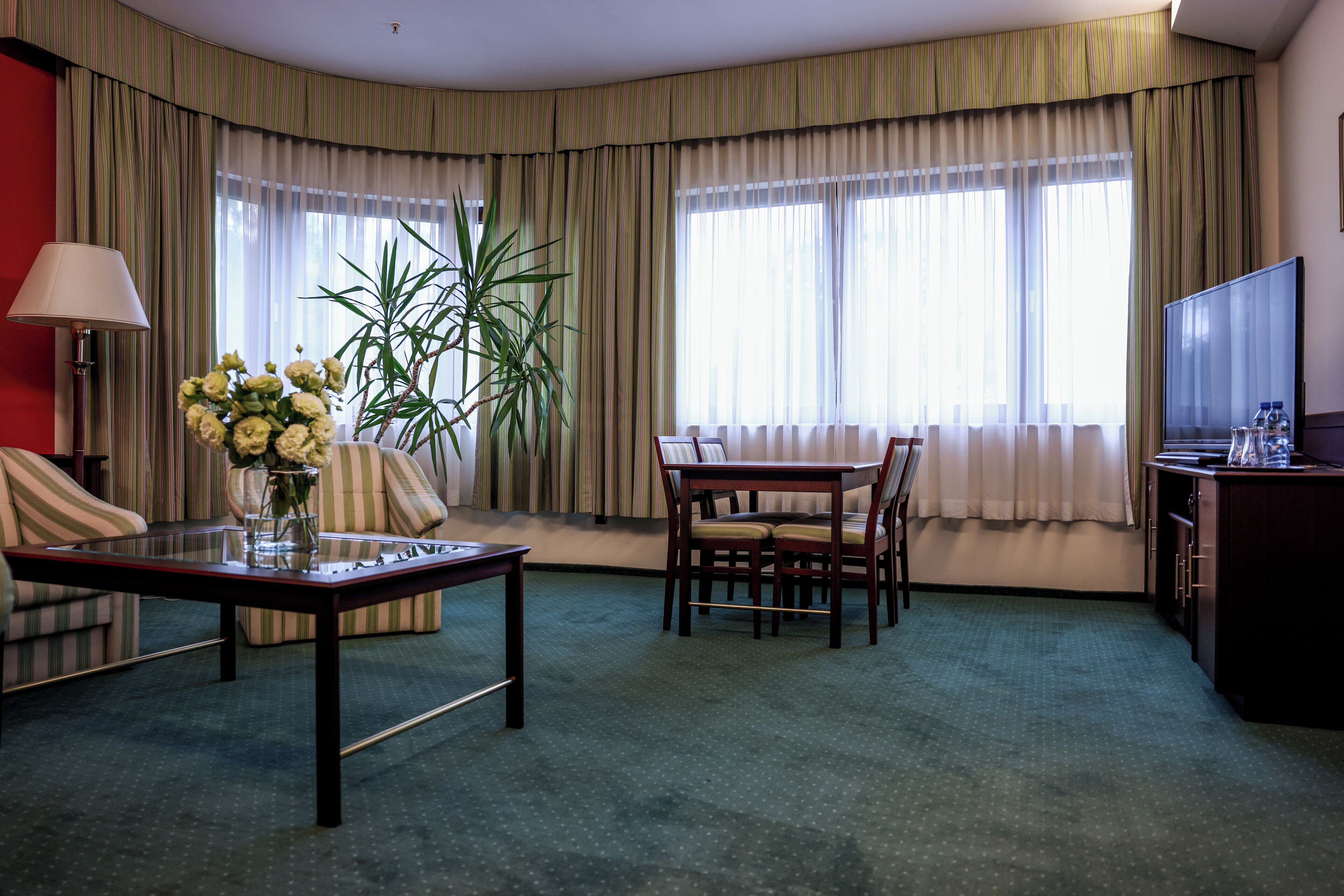 A hotel room with a sofa, a dining table with chairs, a TV, a lamp, and a plant.