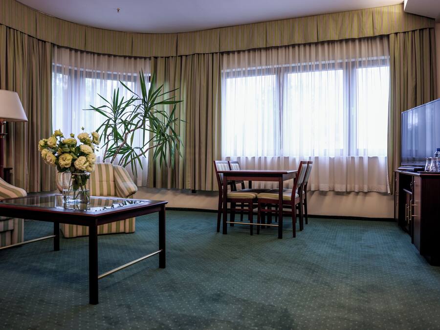 A hotel room with a sofa, a dining table with chairs, a TV, a lamp, and a plant.