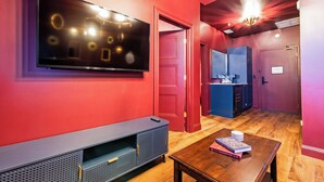 Habitación de hotel moderna con un televisor de pantalla plana grande montado en la pared, una mesa de centro de madera con libros, una pequeña kitchenette y una puerta roja.