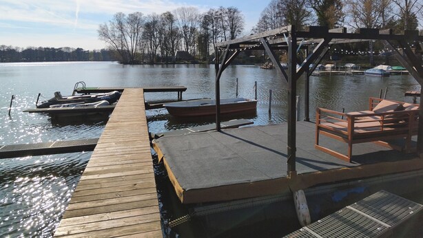 Une jetée en bois s’étendant sur un lac paisible, où sont amarrés des bateaux, avec un espace de repos abrité et une vue dégagée sur les arbres environnants.