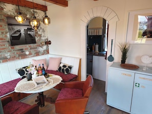 A cozy room with a brick wall, a wooden ceiling, a dining area with a table and chairs, and a kitchen visible through an archway.
