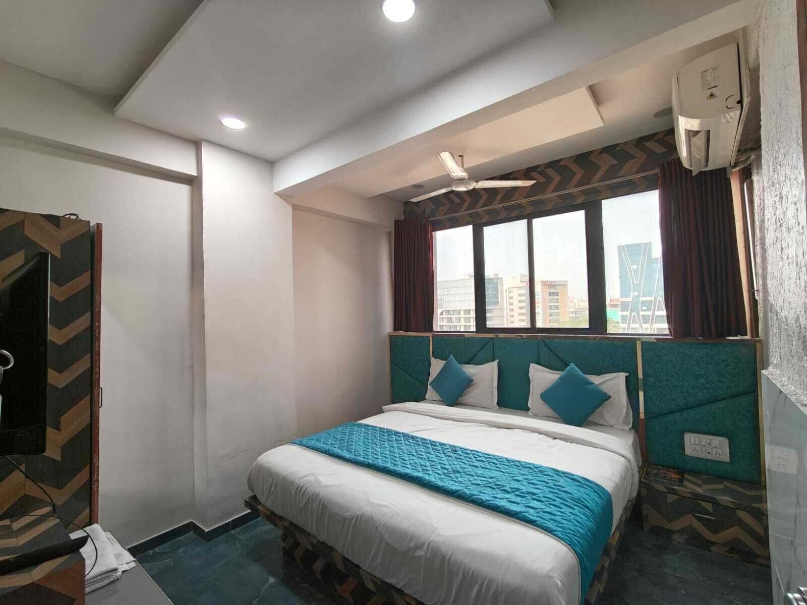 A hotel room with a bed, a ceiling fan, a window with curtains, and a view of buildings outside.