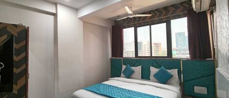 A hotel room with a bed, a ceiling fan, a window with curtains, and a view of buildings outside.
