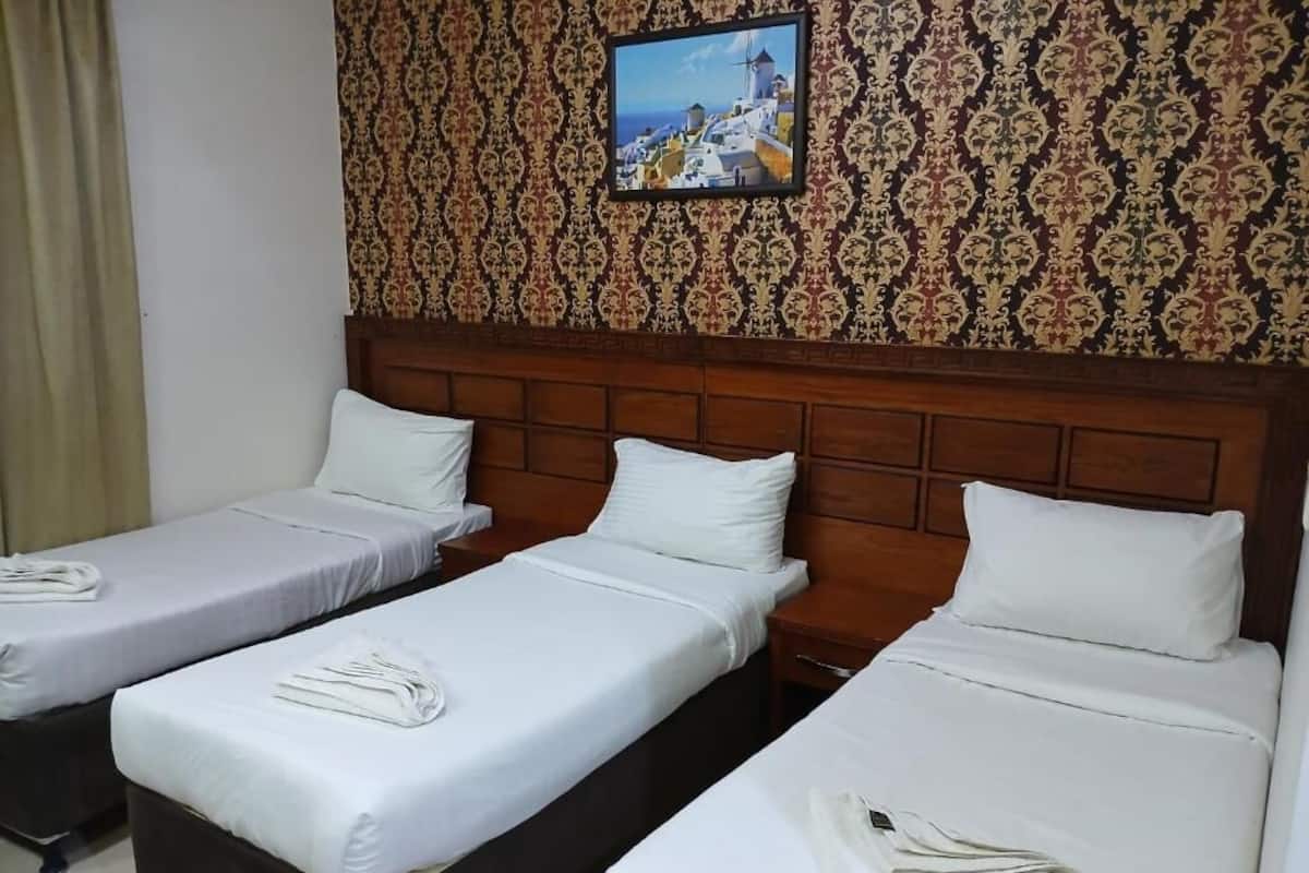A hotel room with three single beds, each with white linens and pillows. The room has a wooden headboard and a patterned wallpaper with a framed picture above the beds.