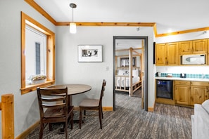 A compact kitchen with wooden cabinets, a dining area with a round table and chairs, and a visible doorway leading to a bedroom with a bunk bed.