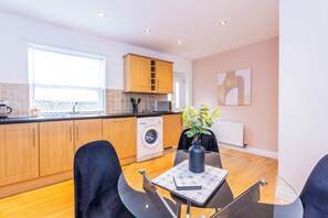 A modern kitchen with wooden cabinets, a washing machine, and a dining area with a glass table and chairs. - Toast Stays - The Peth House (Shildon)