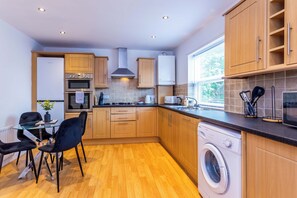 A compact kitchen with wooden cabinets, a built-in oven, a microwave, a washing machine, and a dining area with a glass table and chairs. - Toast Stays - The Peth House (Shildon)