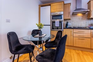 A modern kitchen with a glass dining table, black chairs, a refrigerator, an oven, and a gas stove. - Toast Stays - The Peth House (Shildon)