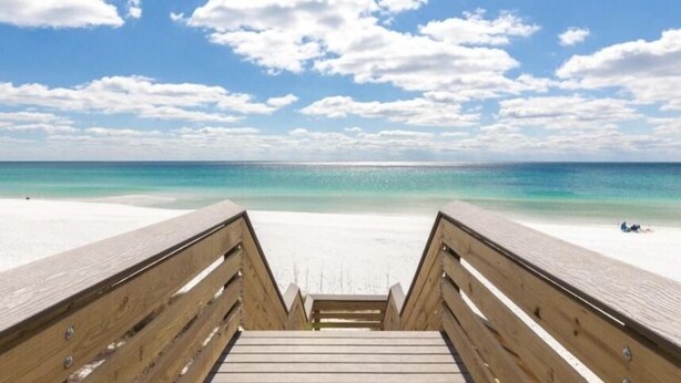 A wooden boardwalk leading to a beach with clear turquoise water and a partly cloudy sky.