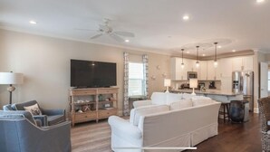 A spacious living room with a large flat-screen TV, a sofa, and a dining area with a kitchen in the background.