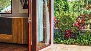 Una casa moderna con interior de madera, un ventanal de vidrio que da a un jardín con plantas tropicales y un cuadro enmarcado en la pared.