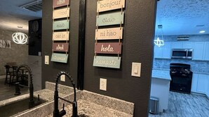 Una cocina moderna con encimeras de granito, un lavamanos de acero inoxidable y un letrero de madera colgante con saludos en varios idiomas.