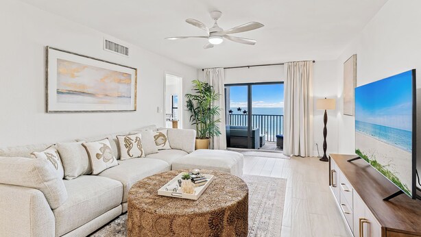 A modern living room with a sofa, coffee table, and a TV. There's a balcony with a view of the sea.