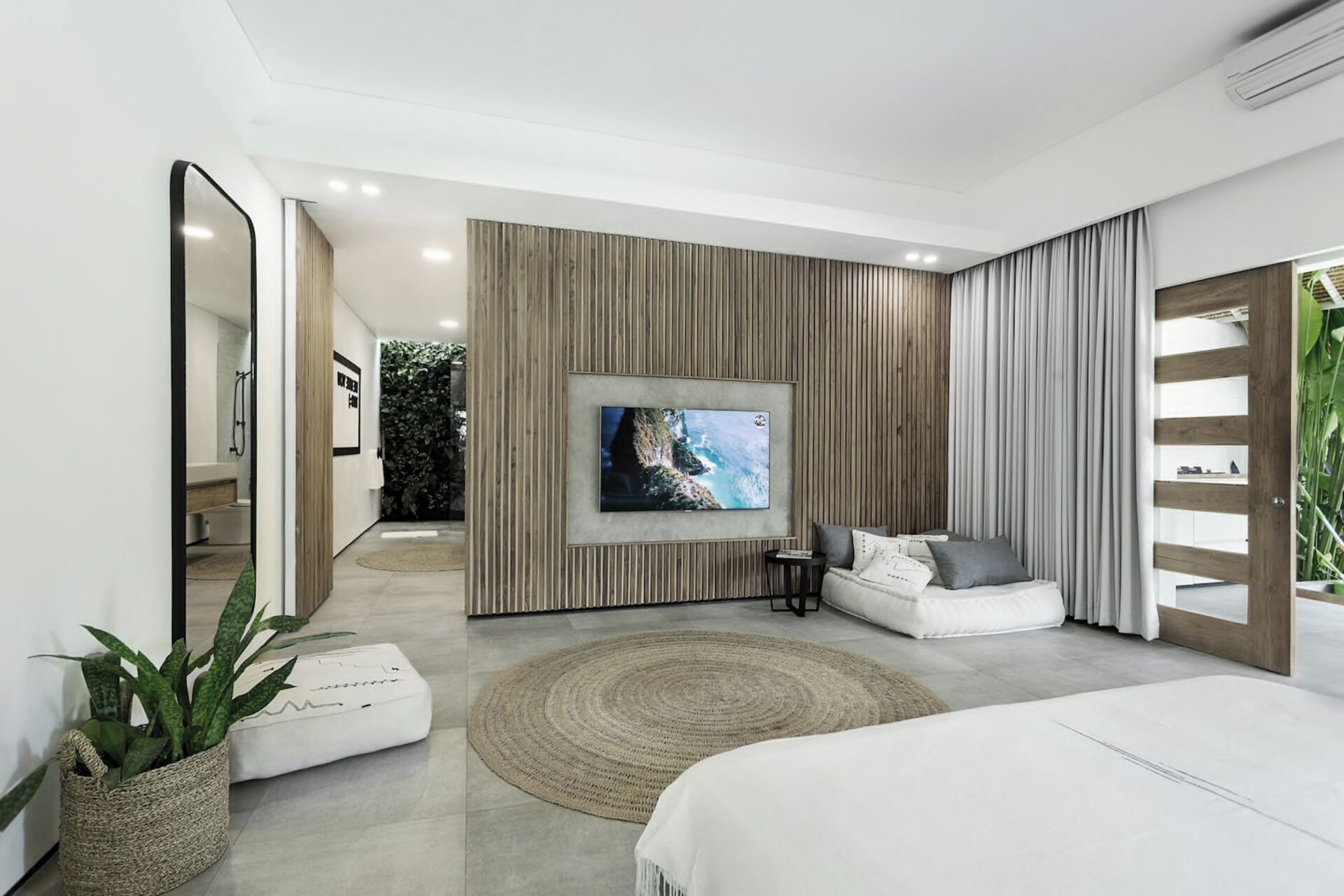 A modern bedroom with a large bed, a TV mounted on a wooden panel wall, a seating area with a sofa and cushions, and a large mirror.