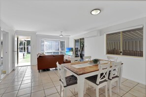 A bright living room with a sofa, a dining table with chairs, and a TV. There is a sliding glass door leading to a balcony. - The location couldn’t be more convenient (Amity)