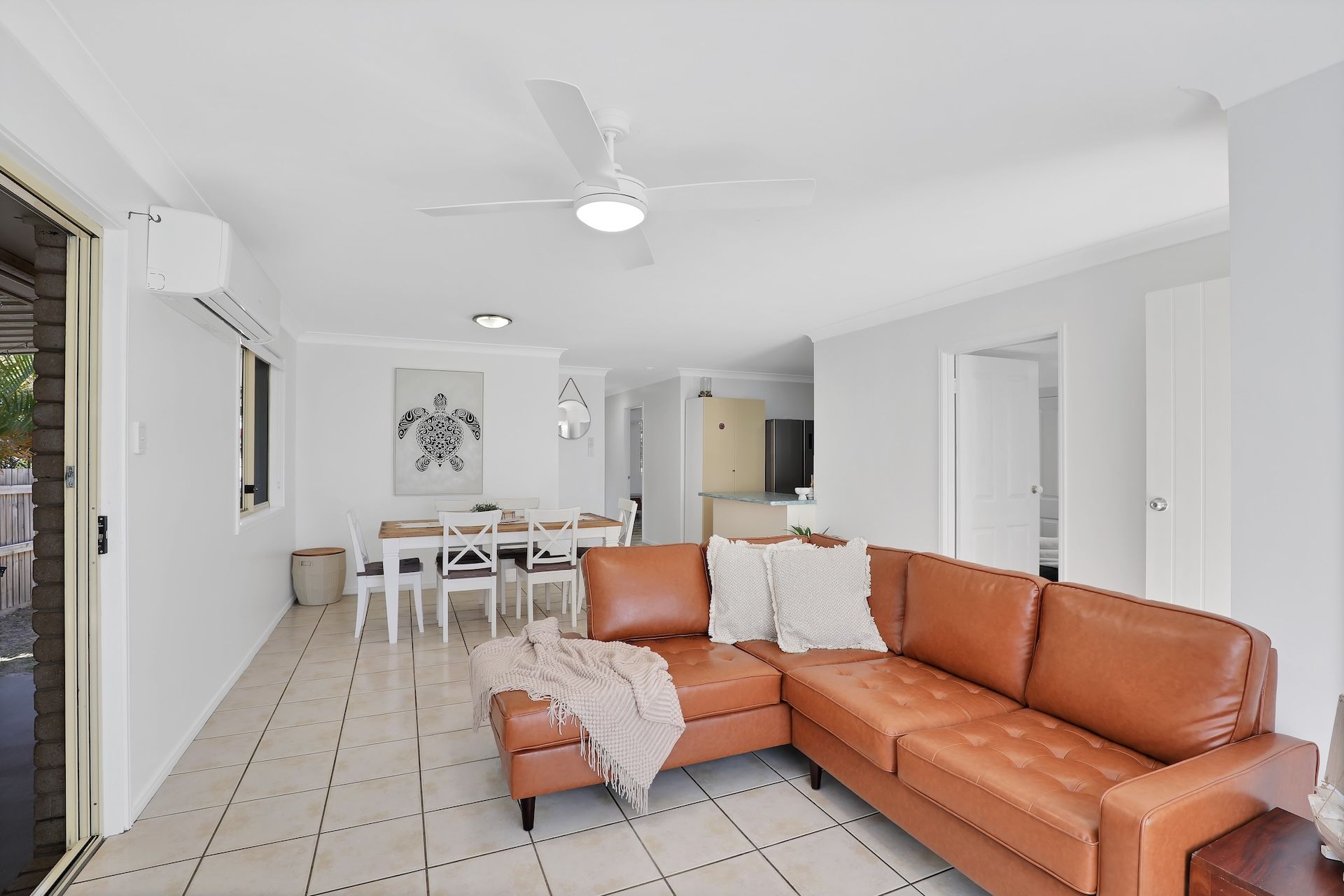 Un salon avec un canapé en cuir marron, une table à manger avec des chaises, un ventilateur de plafond et, au fond, un coin cuisine.