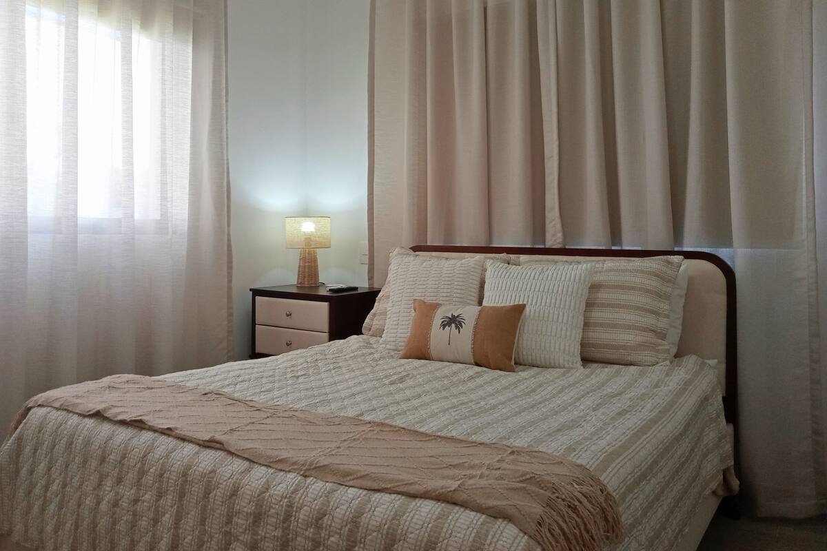 A neatly made bed with a patterned bedspread and a throw blanket, a bedside table with a lamp, and sheer curtains.