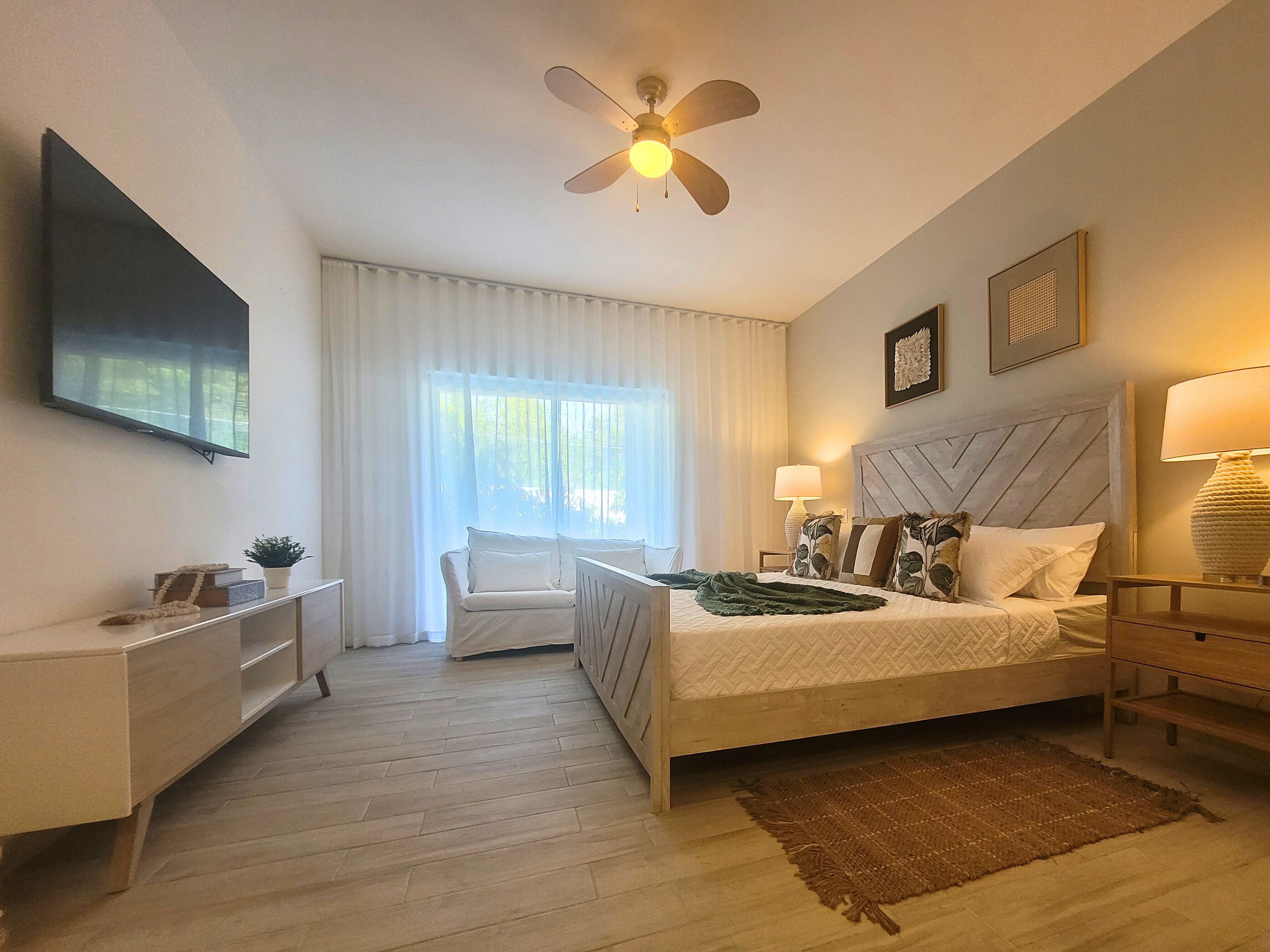 A bedroom with a bed, bedside tables, a chair, a ceiling fan, a TV, and a window with curtains.
