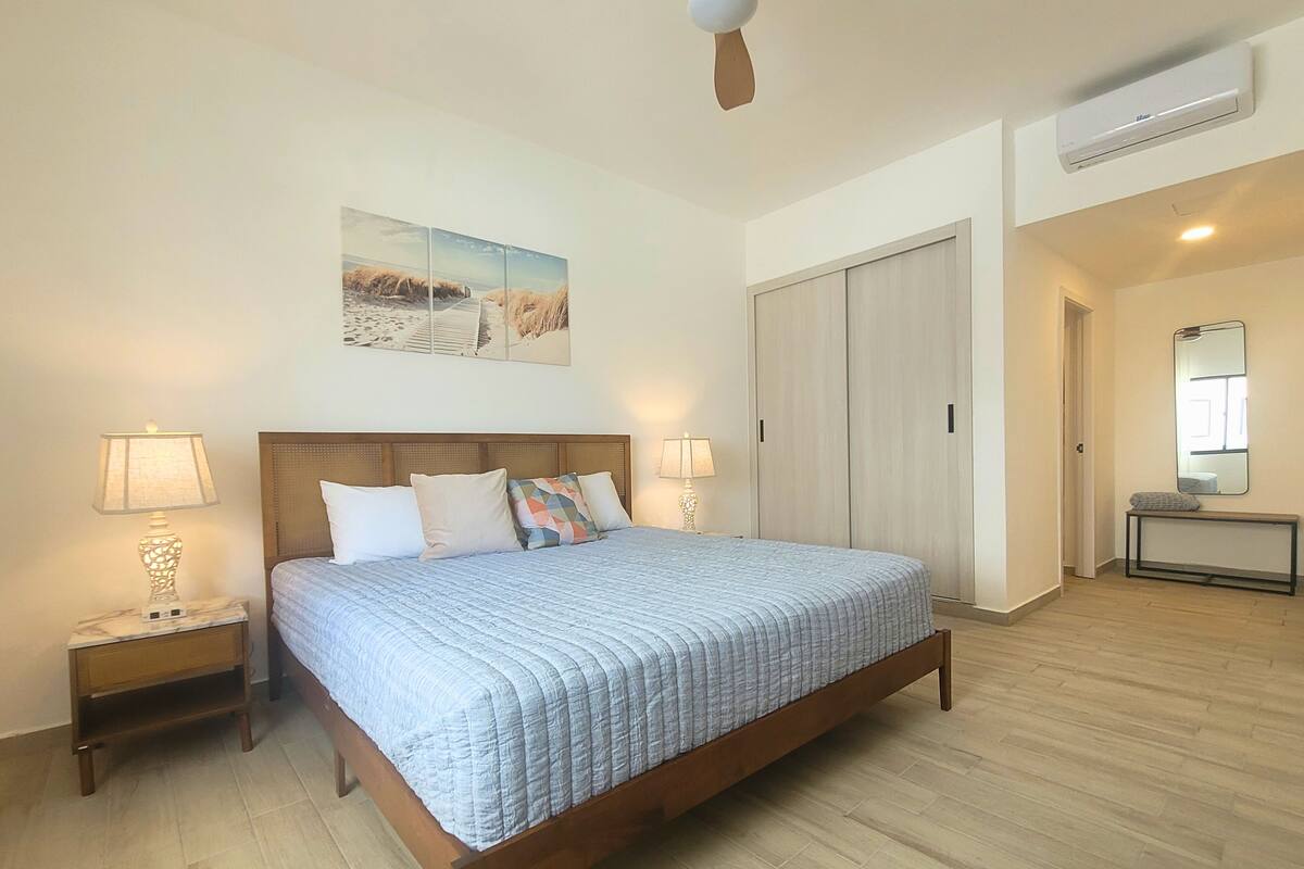 A bedroom with a wooden bed, two bedside lamps, a ceiling fan, an air conditioning unit, a large mirror, and a picture on the wall.