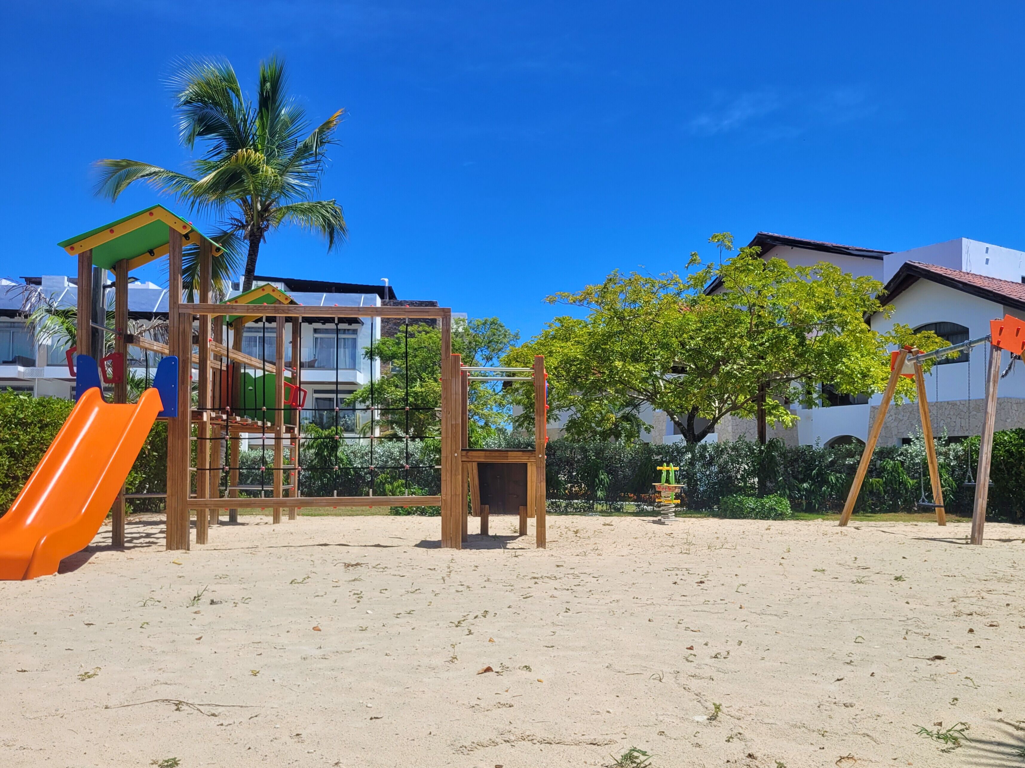 A playground with a slide and climbing structures, surrounded by sand and palm trees, with buildings in the background.
