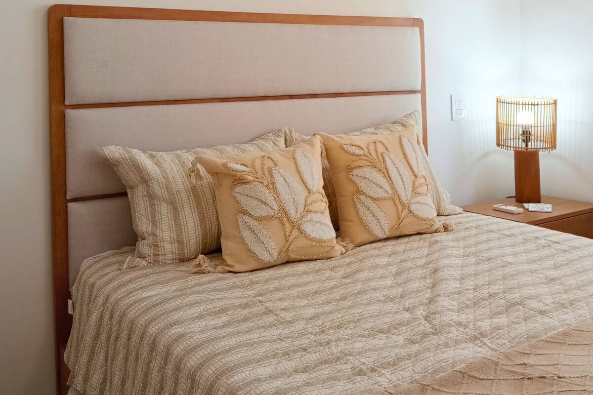 A bed with a headboard, two pillows, a bedside table with a lamp, and a wall-mounted macrame piece.