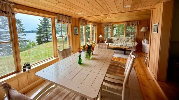 A wooden cabin interior with a dining table, chairs, and a sofa. Large windows offer views of a lake and forest.