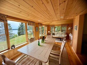 A wooden cabin interior with a dining table, chairs, and a sofa. Large windows offer views of a lake and forest.