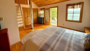 A bedroom with a wooden ceiling, a bed with a patterned bedspread, a window with curtains, and a view of greenery outside.