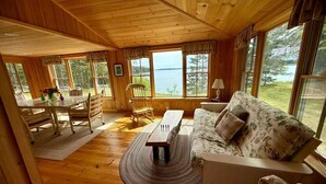 A wooden cabin interior with a dining area, a sofa, and large windows overlooking a body of water.