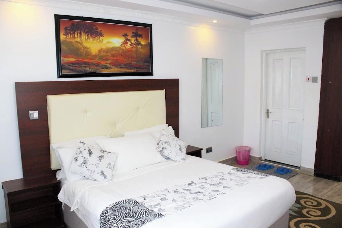 A hotel room with a large bed, a nightstand, a painting above the bed, a door, and a small trash bin.