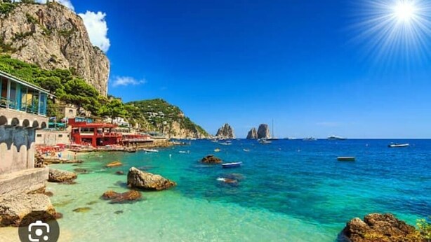 A coastal scene with clear turquoise waters, rocky outcrops, and a building with a red facade.