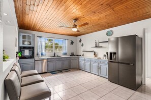 A kitchen with a wooden ceiling, a refrigerator, cabinets, a sink, and a window with curtains.