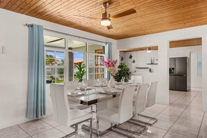 A modern dining area with a glass table and white chairs, a ceiling fan, and a view of the outdoors. - Incredible 4 BR Home with Private Pool Near Ocean (Pos Chiquito)