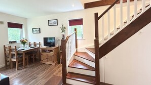 A dining area with a wooden table and chairs, a television, and a cabinet. There is a staircase with wooden steps and white railings.